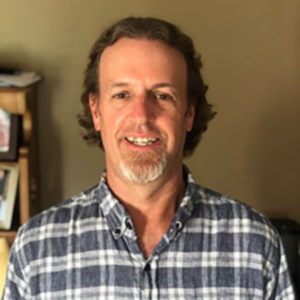 UCLA Professor Burt Cowgill wearing plaid grey button up shirt against a tan background, indoors