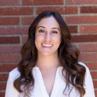 A headshot of Jenna Mendoza, UCLA MHA Student Affairs Officer, smiling warmly in front of a red brick wall. She has long, wavy brunette hair and is wearing a white, short-sleeved textured knit top with three large brown buttons down the front. The lighting is bright and natural, creating an approachable and professional appearance.