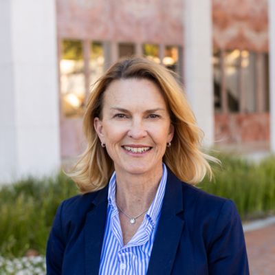 Dr. Leah Vriesman stands outdoors against a red brick building, wearing a navy blazer and blue and white collared shirt