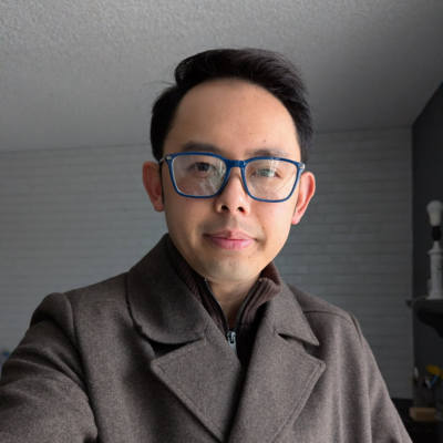 A headshot of Christopher K. Lee, MPH, a lecturer for the UCLA Master of Healthcare Administration (MHA) program. He is a man with short dark hair and blue-rimmed glasses, wearing a brown pea coat over a dark zip-up sweater. He is looking directly at the camera with a neutral, friendly expression against a background of a white brick wall.