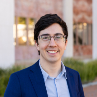 Outdoor headshot portrait of Alan Rozet, a 2025 UCLA EMPH graduate, wearing glasses and smiling warmly at the camera. He is wearing a navy blue blazer over an open-collared light blue dress shirt, with a softly blurred UCLA campus building and greenery in the background.