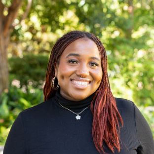 EMPH Student Alexa Osime stands outside near a green-leafed tree. She wears a black turtleneck shirt and a silver necklace.
