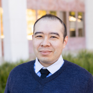 Outdoor headshot portrait of Alvin Wong, a 2025 UCLA EMPH graduate, with short dark hair, looking toward the camera with a gentle smile. He is wearing a dark navy v-neck sweater over a light blue collared dress shirt and dark tie, with a softly blurred UCLA campus building and greenery in the background.