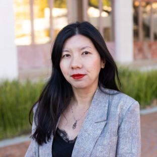 Outdoor headshot portrait of Daisy Wang, a 2025 UCLA EMPH graduate, with long dark hair and red lipstick, facing the camera with a subtle smile. She is wearing a light grey textured blazer over a black lace-trimmed top and a pendant necklace, set against a softly blurred UCLA campus background.