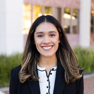 Outdoor headshot portrait of Dayanara Nieto Flores, a 2025 UCLA EMPH graduate, with long dark hair featuring subtle lighter highlights, smiling broadly at the camera. She is wearing a black blazer over a white ribbed top with black trim and buttons, set against a softly blurred UCLA campus background.