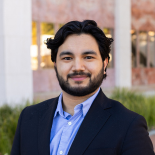 Outdoor headshot portrait of Elias Rodriguez, a 2025 UCLA EMPH graduate, with dark wavy hair and a trimmed beard, smiling warmly at the camera. He is wearing a dark suit jacket over an open-collared light blue shirt, set against a softly blurred UCLA campus background.
