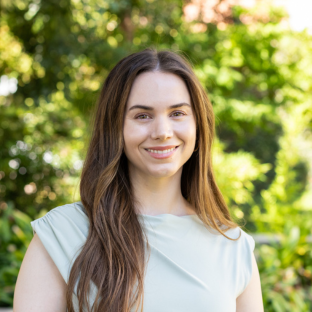 EMPH Student Emily Brown stands outside near a green-leafed tree on the UCLA campus. She wears a light blue top. Her hair is brown, straight and long and parted down the center.