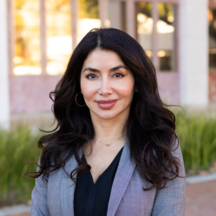 Outdoor headshot portrait of Eunice Ninet, a 2025 UCLA EMPH graduate, with long, wavy dark hair and gold hoop earrings, smiling gently at the camera. She is wearing a grey textured blazer over a black v-neck top, set against a softly blurred UCLA campus background with greenery.