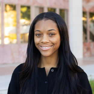 Outdoor headshot portrait of Kennedy Anderson, a 2026 UCLA EMPH graduate, with long, straight dark hair and light brown eyes, smiling warmly at the camera. She is wearing a black button-down top, set against a softly blurred UCLA campus building in the background.