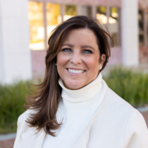 Outdoor headshot portrait of Lisa Edwards, a 2025 UCLA EMPH graduate, with side-swept brown hair and blue eyes, smiling warmly at the camera. She is wearing a cream-colored turtleneck sweater and matching light jacket, set against a softly blurred UCLA campus background.