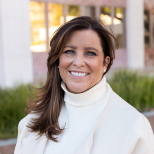 Outdoor headshot portrait of Lisa Edwards, a 2025 UCLA EMPH graduate, with side-swept brown hair and blue eyes, smiling warmly at the camera. She is wearing a cream-colored turtleneck sweater and matching light jacket, set against a softly blurred UCLA campus background.