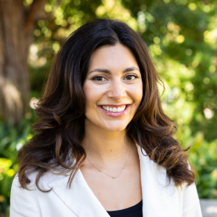 EMPH Candidate stands outside wearing a white blazer over a black top. She stands near a green-leafed tree on the UCLA campus. Her brown hair is shoulder-length and parted down the center.