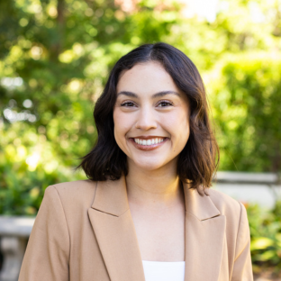 EMPH Candidate Maya Camacho wears a light tan blazer over a white shirt. She stands outside on the UCLA campus near a green-leafed tree. Her short brown hair is parted down the center.