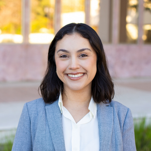 headshot of UCLA Executive MPH graduate Maya Camacho. She has shoulder length dark brown hair and she wears a light blue blazer over a white button-up collared shirt.