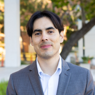 Outdoor headshot portrait of Miguel Ramirez, a 2026 UCLA EMPH graduate, with dark center-parted hair and brown eyes, smiling slightly at the camera. He is wearing a grey plaid suit jacket over an open-collared light dress shirt, with a softly blurred outdoor campus background.