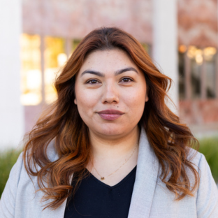 Outdoor headshot portrait of Nicole Morales, a 2025 UCLA EMPH graduate, with wavy reddish-brown hair and dark eyes, smiling gently at the camera. She is wearing a light grey blazer over a black top and a delicate gold necklace, with a softly blurred UCLA campus background.