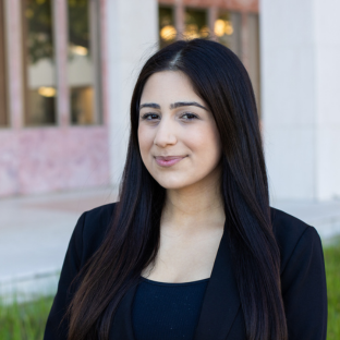 Outdoor headshot portrait of Nicollette Doomany, a 2026 UCLA EMPH graduate, with long, straight dark hair, smiling gently at the camera. She is wearing a black blazer over a dark top, with a softly blurred campus background.