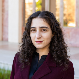 Headshot portrait of Sarah Hakakha, a 2026 UCLA EMPH graduate, with long curly dark hair and brown eyes, smiling at the camera. She is wearing a maroon blazer over a black top, with a softly blurred campus building in the background.