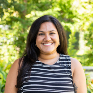EMPH Candidate Trishna Singh stands outside on the UCLA campus wearing a sleeveless black and white top. There is a green leafy tree behind her. Her long dark brown hair is parted on the side and hangs over one shoulder.