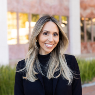 Outdoor headshot portrait of Vanessa Villafuerte, a 2025 UCLA EMPH graduate, with long blonde-highlighted hair and a broad smile, looking at the camera. She is wearing a black blazer over a black turtleneck, set against a softly blurred UCLA campus background with greenery.
