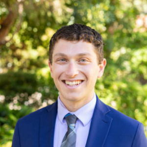 EMPH Candidate Zachary Morganstein wears a royal blue blazer over a lavendar colored shirt and tie. He stands outside on the UCLA campus with a green, leafy tree in the background.