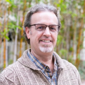 Professor Burt Cowgill stands outside with green shrubbery in the background. Cowgill wears a light brown sweater over a light blue and brown plaid collared button-up shirt.