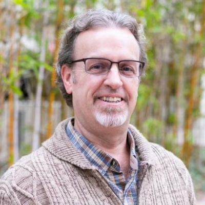 Professor Burt Cowgill stands outside with green shrubbery in the background. Cowgill wears a light brown sweater over a light blue and brown plaid collared button-up shirt.