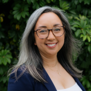 Dr. Isomi Miyake-Lye stands outside in front of a green plant. She wears eyeglasses and a blue blazer over a white top.