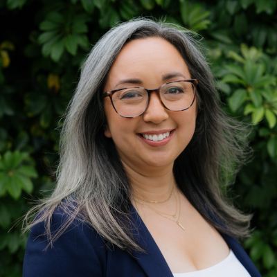 Dr. Isomi Miyake-Lye stands outside in front of a green plant. She wears eyeglasses and a blue blazer over a white top.