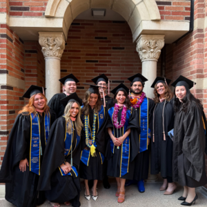 9 EMPH students standing in front of a brick building on the UCLA campus as they wear their graduation cap and gown
