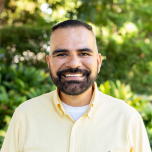 UCLA EMPH Candidate Alfonso Iniguez stands outside on the UCLA campus in front of a green, leafy tree. He is wearing a light yellow collared top, buttoned in the front, over a white t-shirt. He has short dark brown hair, moustache, and beard.