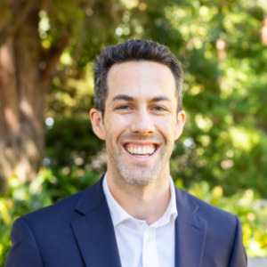 UCLA EMPH Candidate Blake Owens stands outside on the UCLA campus in front of a green, leafy tree. He is wearing a navy blue blazer over a white button-up collared shirt. He has short brown hair and is smiling.