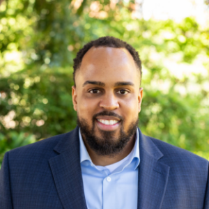 EMPH Candidate Devon Anderson stands outside on the UCLA campus with a green, leafy tree in the background. He wears a navy blue blazer over a light blue colored shirt. He has dark hair, a moustache and a beard.