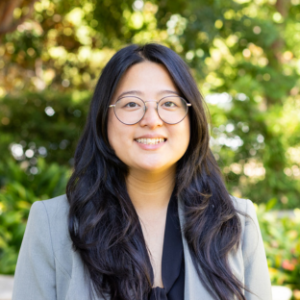 UCLA EMPH Candidate Isabella McShane stands outside on the UCLA campus in front of a green, leafy tree. She is wearing a light grey blazer over a black top. She is wearing thin, wire framed eyeglasses. She has long, straight black hair parted down the center and hanging in front of both shoulders.