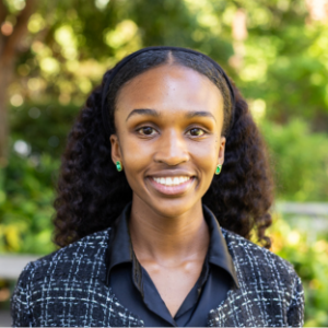 UCLA EMPH Candidate Joelle Ragland stands outside on the UCLA campus in front of a green, leafy tree. She is wearing a black and white plaid boucle collarless blazer over a black collared shirt. Her long wavy black hair is parted down the center. She is wearing small green earrings.