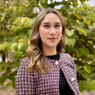 EMPH Student Affairs Office Johanna Arias standing outside wearing a plaid blazer with a wall of leafy green shrubs behind her