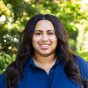 UCLA EMPH Candidate Kisa Hilliard stands outside on the UCLA campus in front of a green, leafy tree. She is wearing a royal blue collared top, buttoned in the front. Her dark brown, long hair is parted down the center and hangs in front of her shoulders.