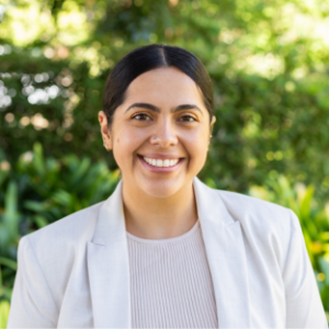 UCLA EMPH Candidate Lauren Garcia stands outside on the UCLA campus in front of a green, leafy tree. She is wearing a white blazer over a white shirt. Her dark brown hair is parted down the center and pulled back.