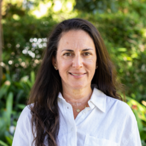 UCLA EMPH Candidate Monique Araya, M.D., stands outside on the UCLA campus, wearing a white long sleeve shirt with a collar and buttons up the front. Her long brown hair is parted on the left side and hangs over one shoulder.