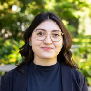 UCLA EMPH Candidate Suini Torres stands outside on the UCLA campus in front of a green, leafy tree. She is wearing a dark blue blazer over a navy blue top. Her dark brown, wavy, shoulder-length hair is parted down the center. She wears eyeglasses.