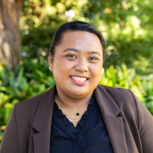 UCLA EMPH Candidate Veronica Tayo stands outside on the UCLA campus in front of a green, leafy tree. She is wearing a brown blazer over a navy blue top. Her dark brown hair is pulled back from her face. She is smiling.