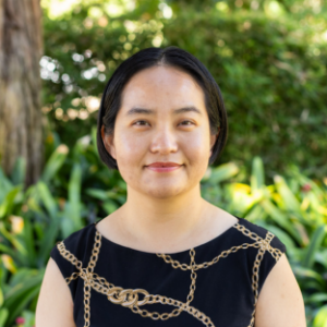UCLA EMPH Candidate Wei Zhao stands outside on the UCLA campus in front of a green, leafy tree. She is wearing a black sleeveless dress with a print of a gold chain. Her short black hair is parted down the center.