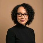 Headshot of UCLA Fielding School of Public Health Executive Master of Public Health Graduate Dr. Cozzette-Lyons Jones as she wears a black turtleneck shirt and black eyeglasses. She stands against a light beige backdrop.