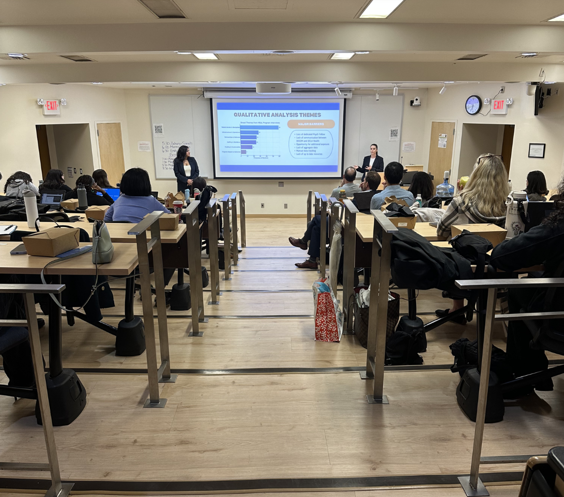 Two UCLA Fielding School of Public Health Executive Master of Public Health (EMPH) second-year students, Trishna Singh and Emily Brown, stand on either side of a projection screen presenting their EMPH capstone project findings to an audience. The slide title reads "QUALITATIVE ANALYSIS THEMES" and displays a bar chart and bullet points. The audience, composed of EMPH cohorts, faculty, and preceptors, is seated in a tiered UCLA lecture hall, with some attendees visible in the foreground working on laptops or listening attentively.