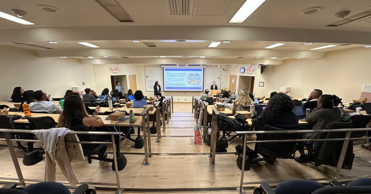 Two UCLA Fielding School of Public Health Executive Master of Public Health (EMPH) second-year students, Trishna Singh and Emily Brown, stand on either side of a projection screen presenting their EMPH capstone project findings to an audience. The audience, composed of EMPH cohorts, faculty, and preceptors, is seated in a tiered UCLA lecture hall, with some attendees visible in the foreground working on laptops or listening attentively.