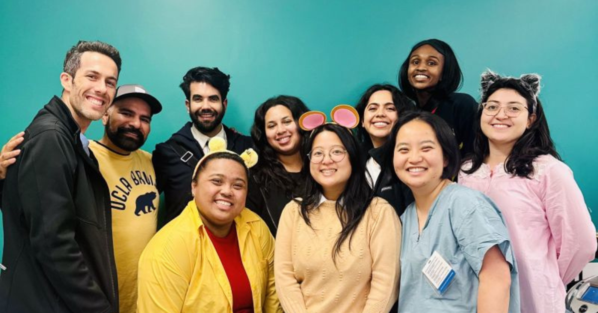 Dr. Dhruv Khurana poses with a group of nine of his UCLA Fielding School of Public Health Executive MPH students in his UCLA class, "Microeconomic Theory of Health Sector," standing close together in front of a teal wall. The group consists of men and women, smiling and looking at the camera. Dr. Dhruv Khurana is the third person from the left in the back row, wearing a dark suit. To the left of him is student Alfonso Iniguez wearing a black and yellow UCLA Bruins t-shirt and a baseball cap. And on the far left is student Blake Owens wearing a dark jacket and smiling. The rest of the group consists of seven female students, some of whom are wearing Halloween costumes or novelty headbands. Student Veronica Tayo, in the front, is wearing a bright yellow shirt, another woman in the center, Isabelle McShane, has pink circular mouse ears, and a woman on the far right, Suini Torres, has grey cat ears. Wei Zhao is wearing light blue medical scrubs. The overall mood is joyful and collegial, reflecting a group of students and their instructor.