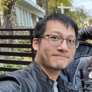 A close-up, outdoor photo of Rui Zhao, an EMPH student, smiling slightly at the camera. He is wearing rectangular metal-framed glasses, a black faux-leather jacket over a grey shirt, and has a short goatee. A black metal fence or railing and some foliage are visible behind him, with the blurred background including a white post and part of a building.