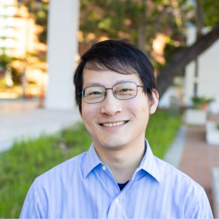 Outdoor headshot portrait of Rui Zhao, a 2026 UCLA EMPH graduate, wearing thin-framed glasses and smiling broadly at the camera. He is wearing a light blue striped button-down shirt, with a softly blurred UCLA campus background featuring greenery and university buildings.