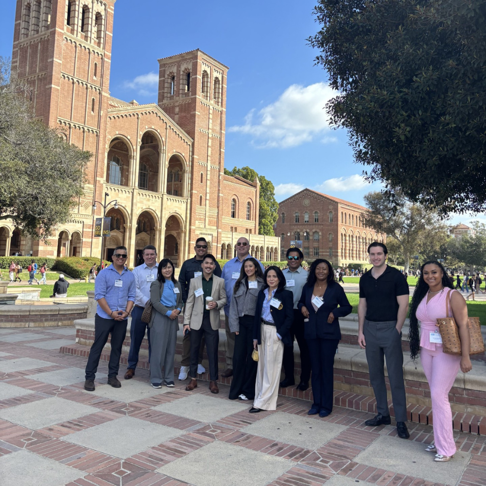 A diverse group of twelve professionally dressed men and women UCLA MHA candidates, many wearing UCLA MHA name tags, pose for a group photo on the UCLA campus. They are standing on a brick landing with the iconic twin-towered, Romanesque-style UCLA Royce Hall and a clear blue sky in the background.