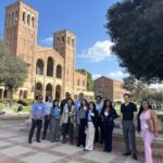 A diverse group of twelve professionally dressed men and women UCLA MHA candidates, many wearing UCLA MHA name tags, pose for a group photo on the UCLA campus. They are standing on a brick landing with the iconic twin-towered, Romanesque-style Royce Hall and a clear blue sky in the background.
